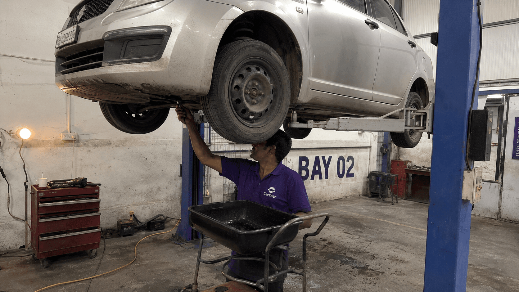 CarYaar technician reaching up to remove the oil drain plug under a car on the workshop lift, with a black drain pan ready below.