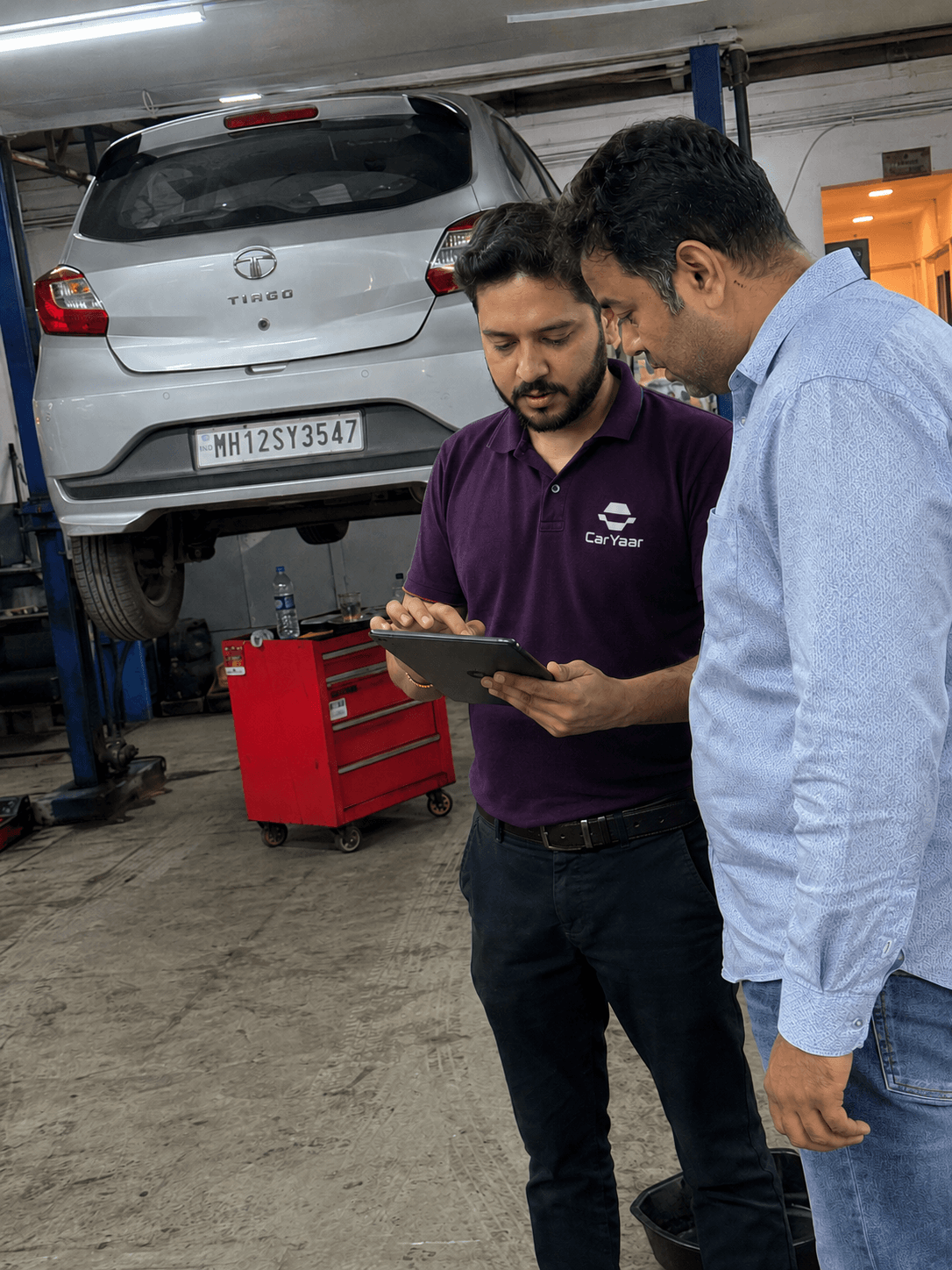 CarYaar service advisor and customer reviewing a service plan on a tablet next to a car on a workshop lift.