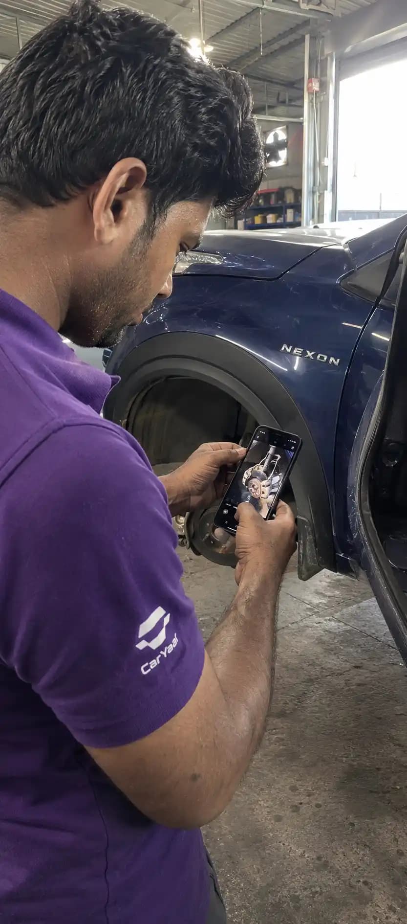 CarYaar technician photographing the brake assembly inside a wheel arch with a phone, for the customer's WhatsApp update.